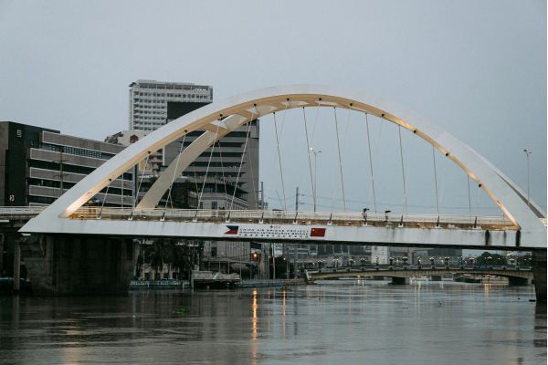 A rusted steel bridge spans a river, with a city skyline visible in the background under a clear blue sky.